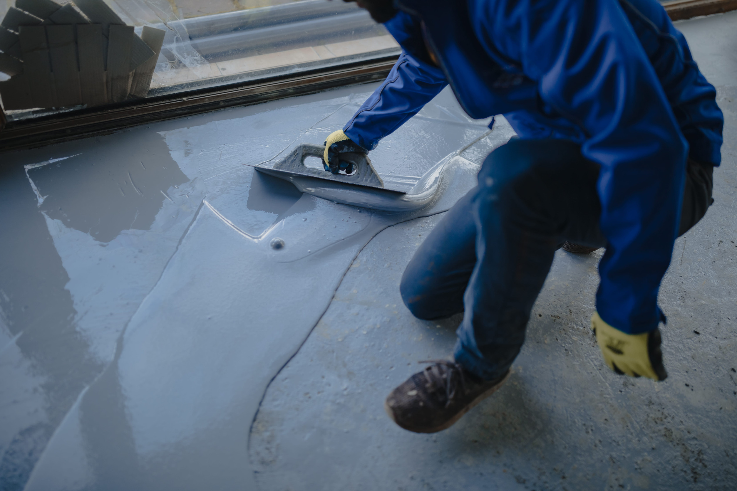 Construction worker in blue coveralls smoothing gray sealant around a roof window with a trowel.