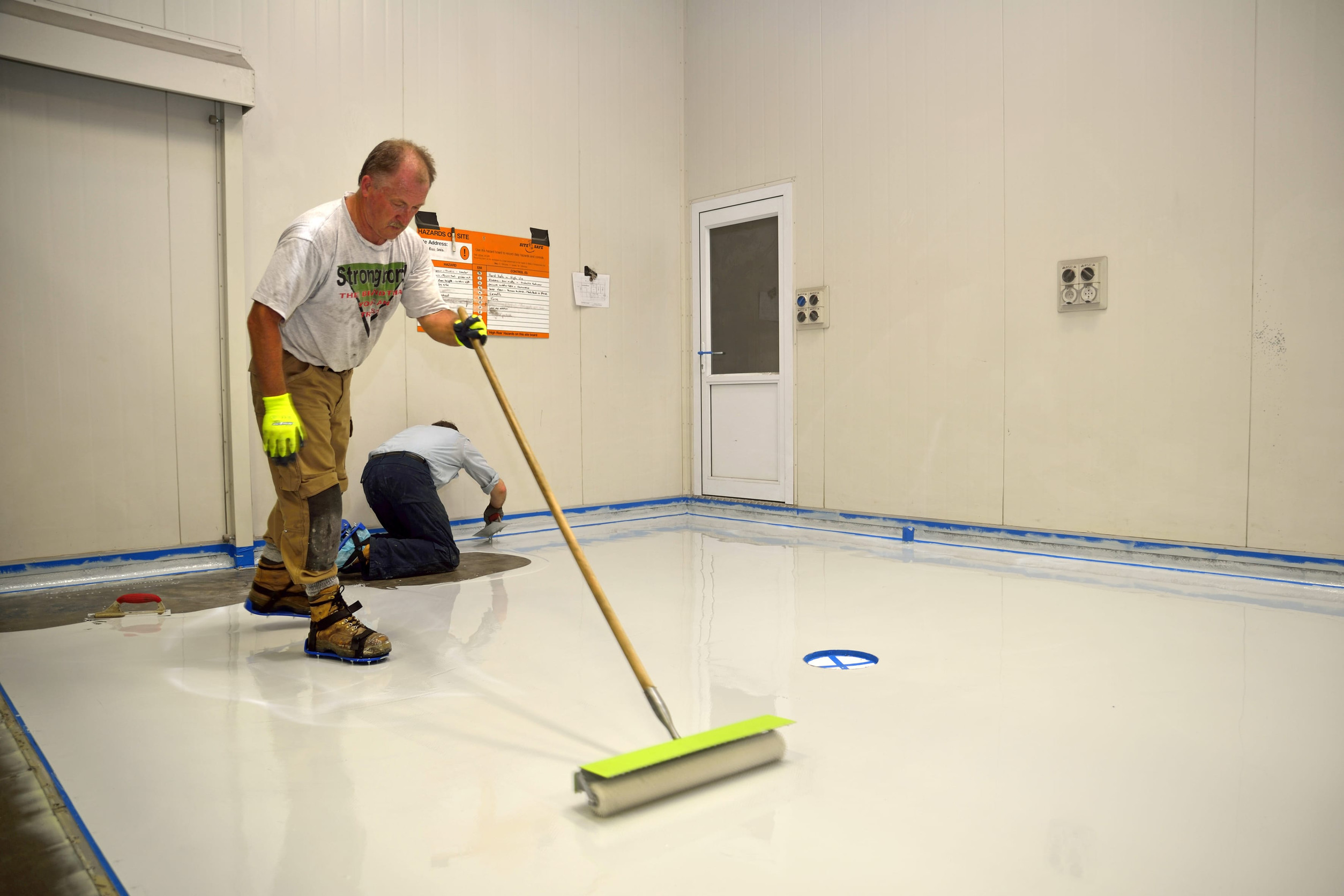 Two workers apply a smooth coat of epoxy garage flooring in a clean, white-walled room; one uses a roller while the other works on the floor near the wall, ensuring long-lasting garage floor durability.