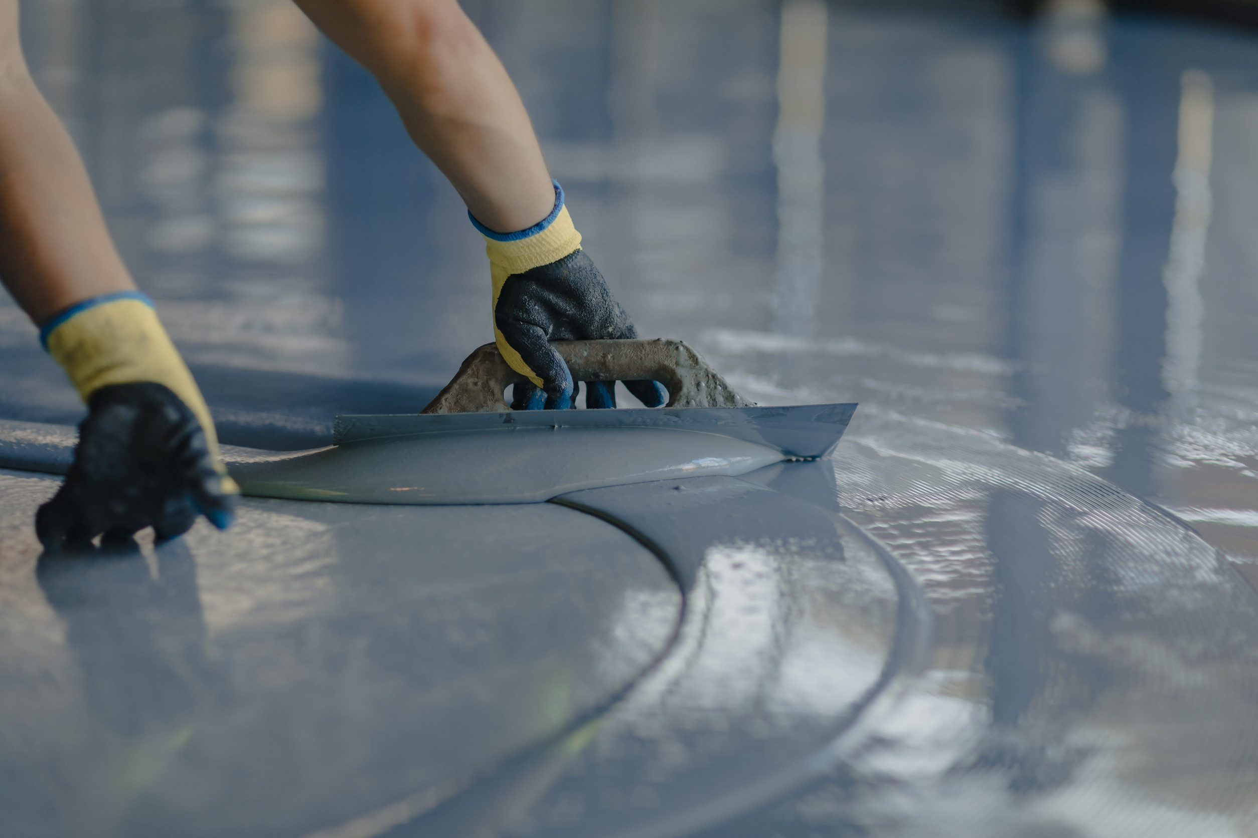A person wearing gloves spreads a layer of epoxy resin onto a floor using a trowel.