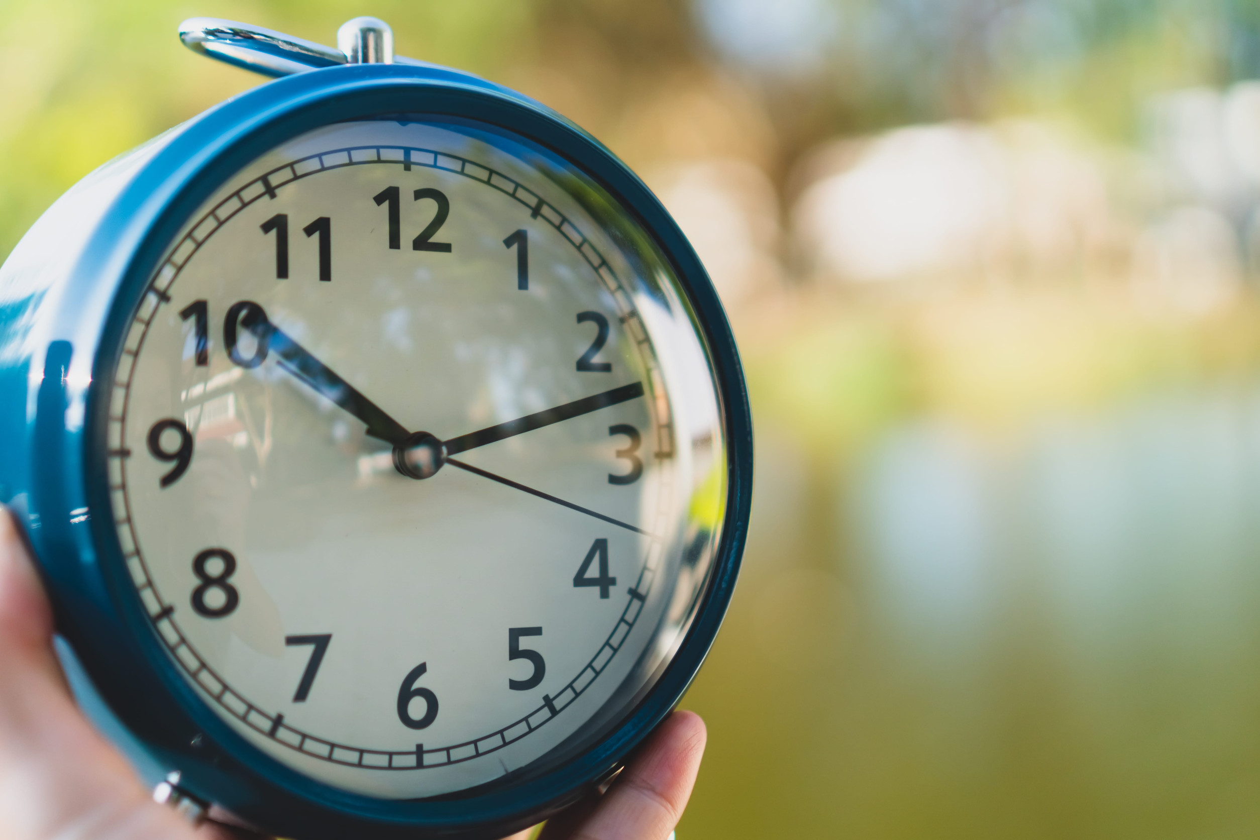 Close-up of a blue alarm clock held in a hand, the clock face showing around 10:10 with a blurred outdoor background.