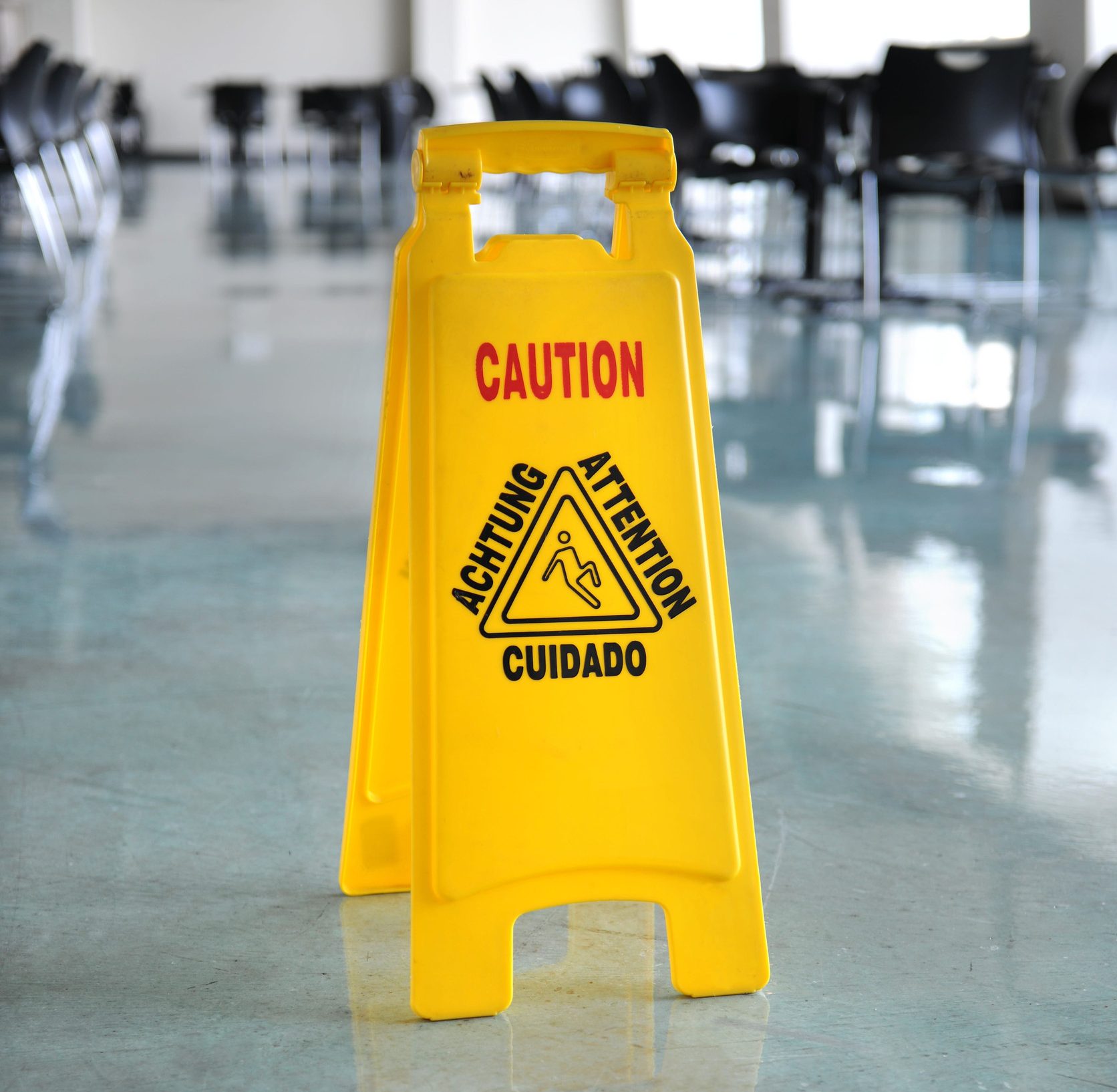 Yellow caution wet floor sign on a shiny, reflective surface in a large room with chairs in the background—a reminder of what you should know about slippery conditions, especially on epoxy garage floors.
