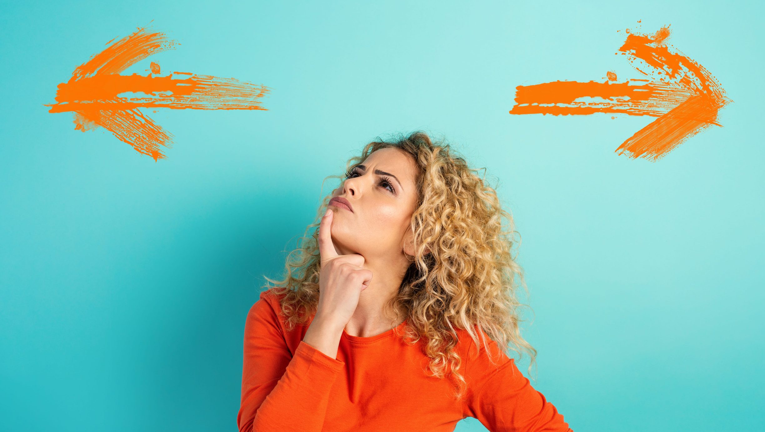 Woman with curly hair in an orange shirt stands against a blue background, looking thoughtful, with orange arrows pointing left and right above her head—perhaps considering the best epoxy garage floor color for her next project.