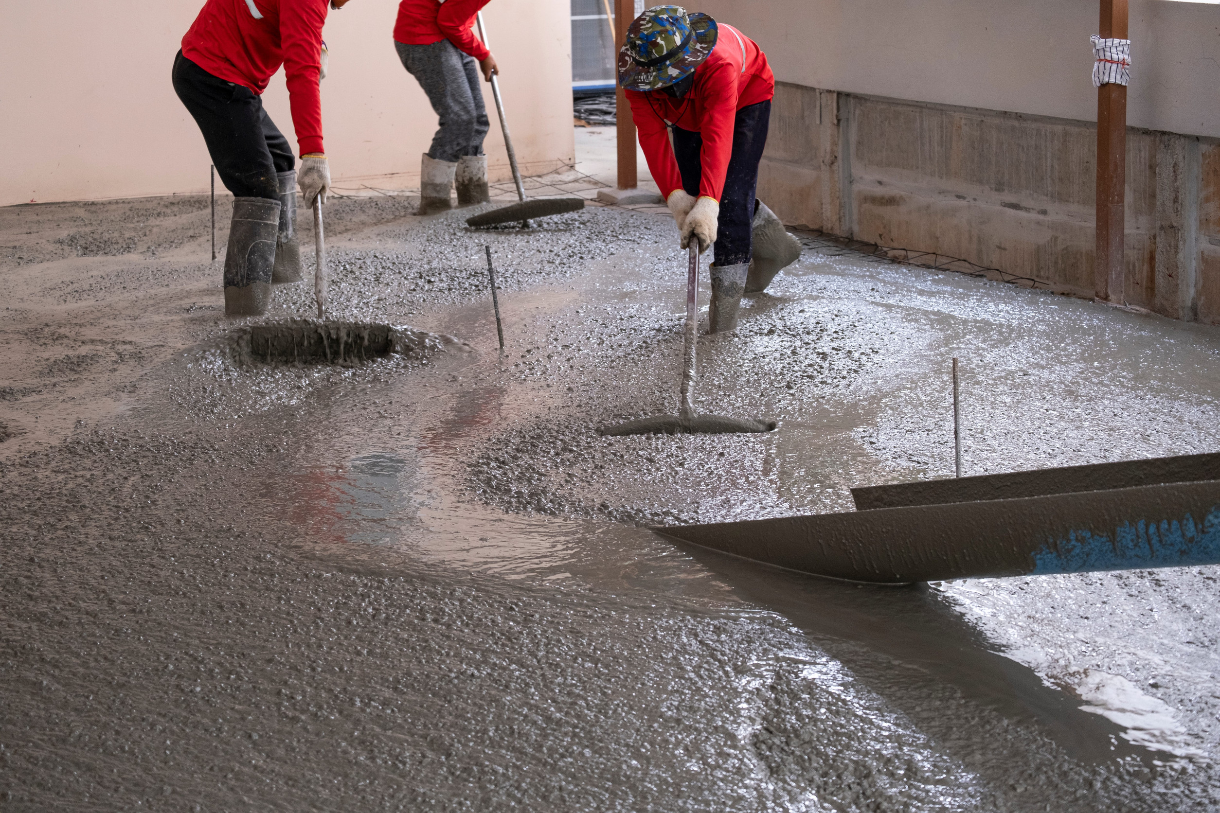 Three workers in red shirts and boots level new concrete on a floor with long tools inside a building under construction, ensuring the surface is prepared for proper concrete cure time before potential installation of epoxy flooring.