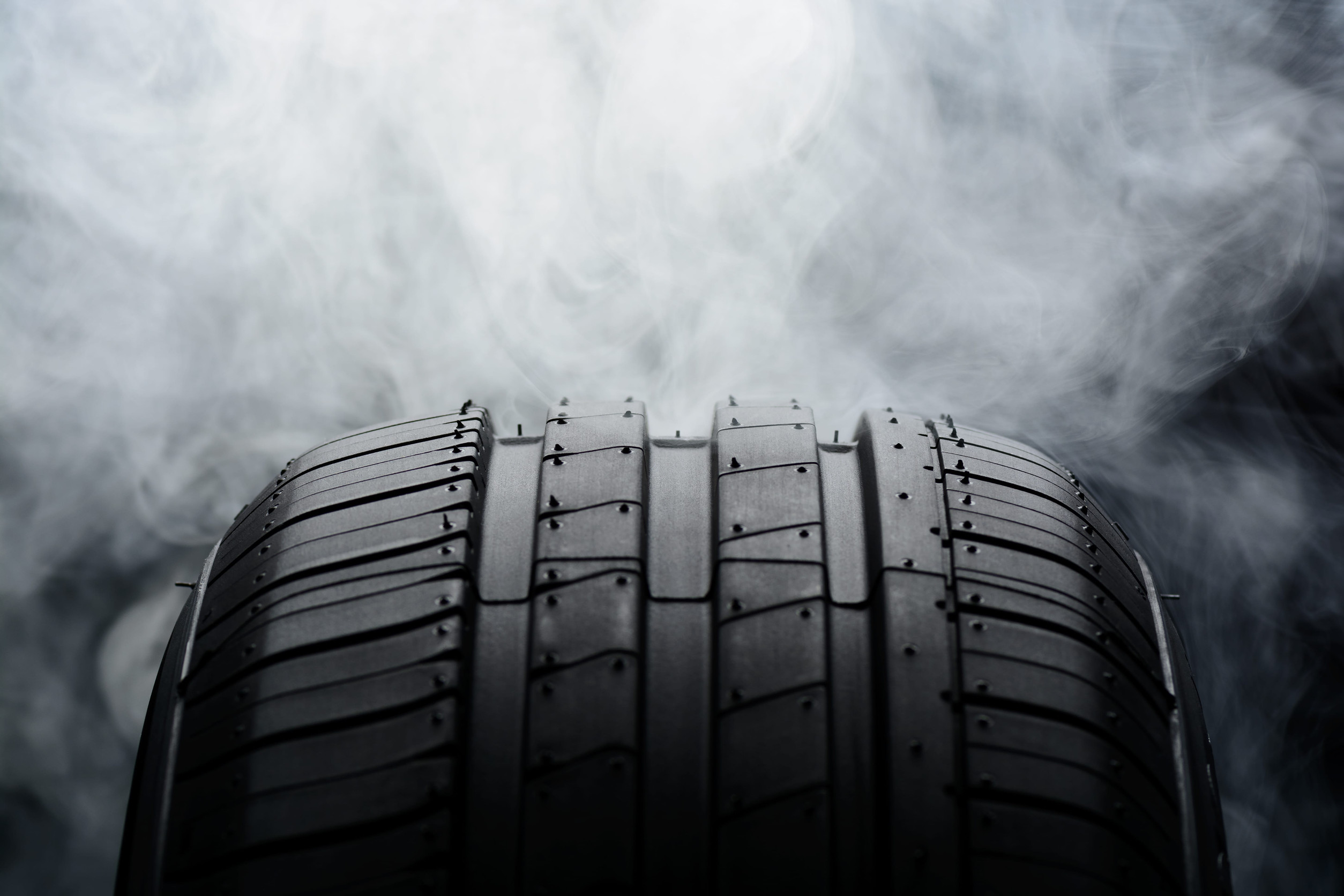Close-up of a car tire tread with wide circumferential grooves, set against a smoky, blurred background.