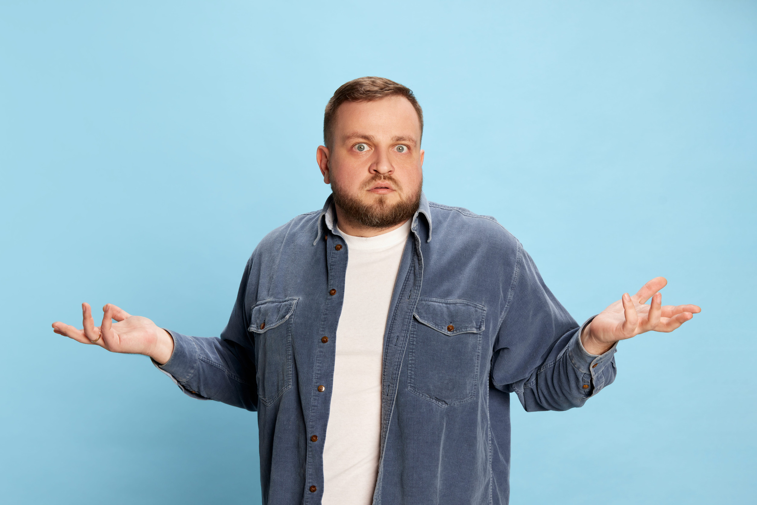 A man in a denim shirt and white t-shirt stands against a blue background, raising his hands and shoulders in a confused or unsure gesture—perhaps illustrating the difference between garage floor paint and epoxy garage flooring.