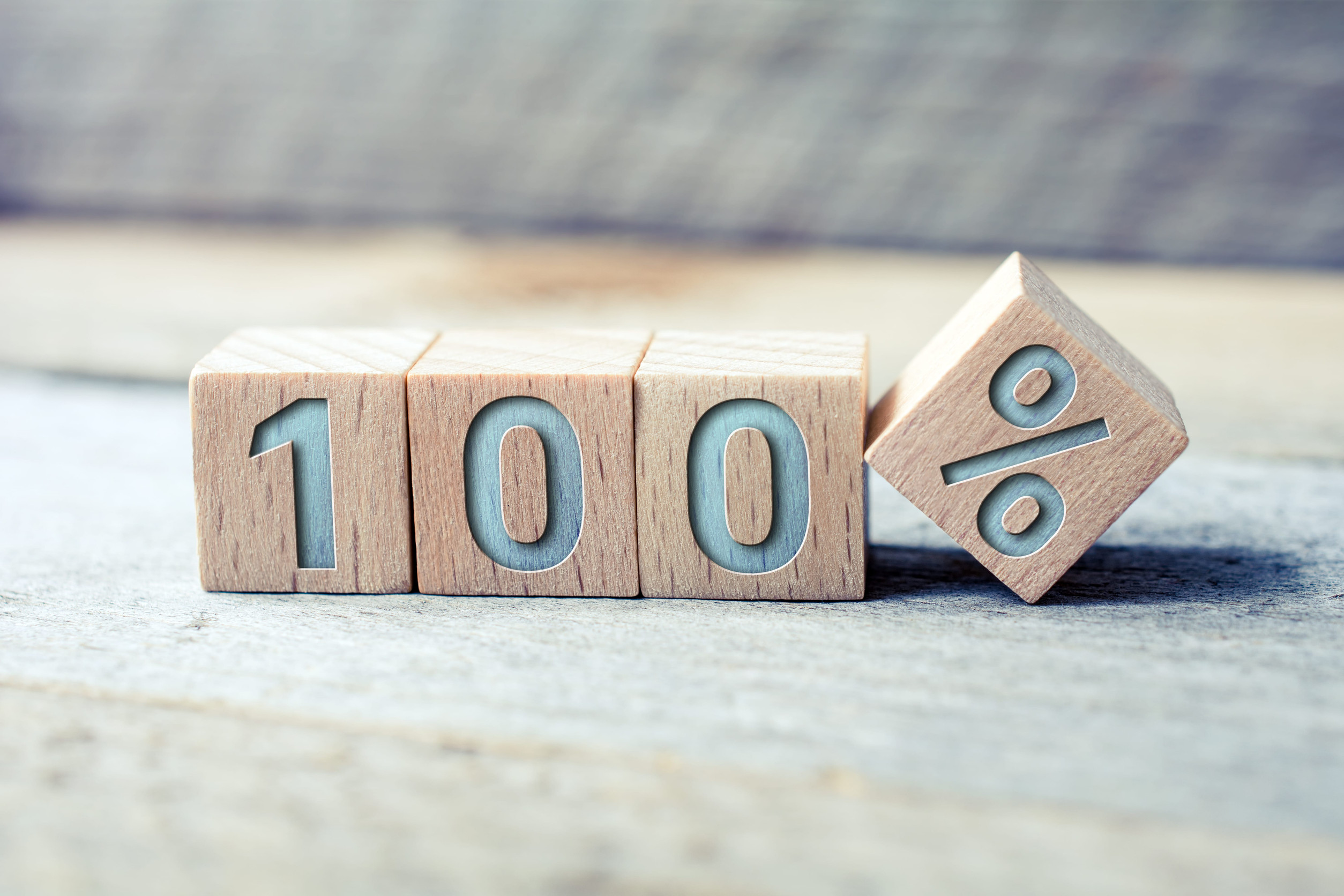 Wooden blocks display “100” with a tilted block showing a percentage sign, placed on a wooden surface reminiscent of a garage floor, evoking the durability and strength of 100% solids epoxy flooring amidst a blurred background.
